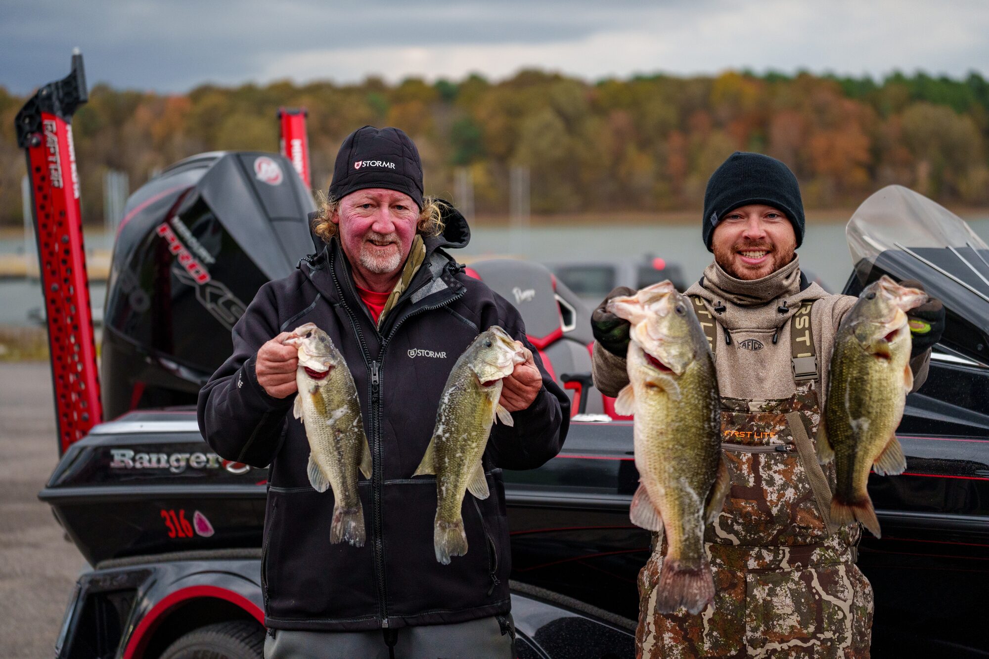Two anglers holding bass beside a tournament boat